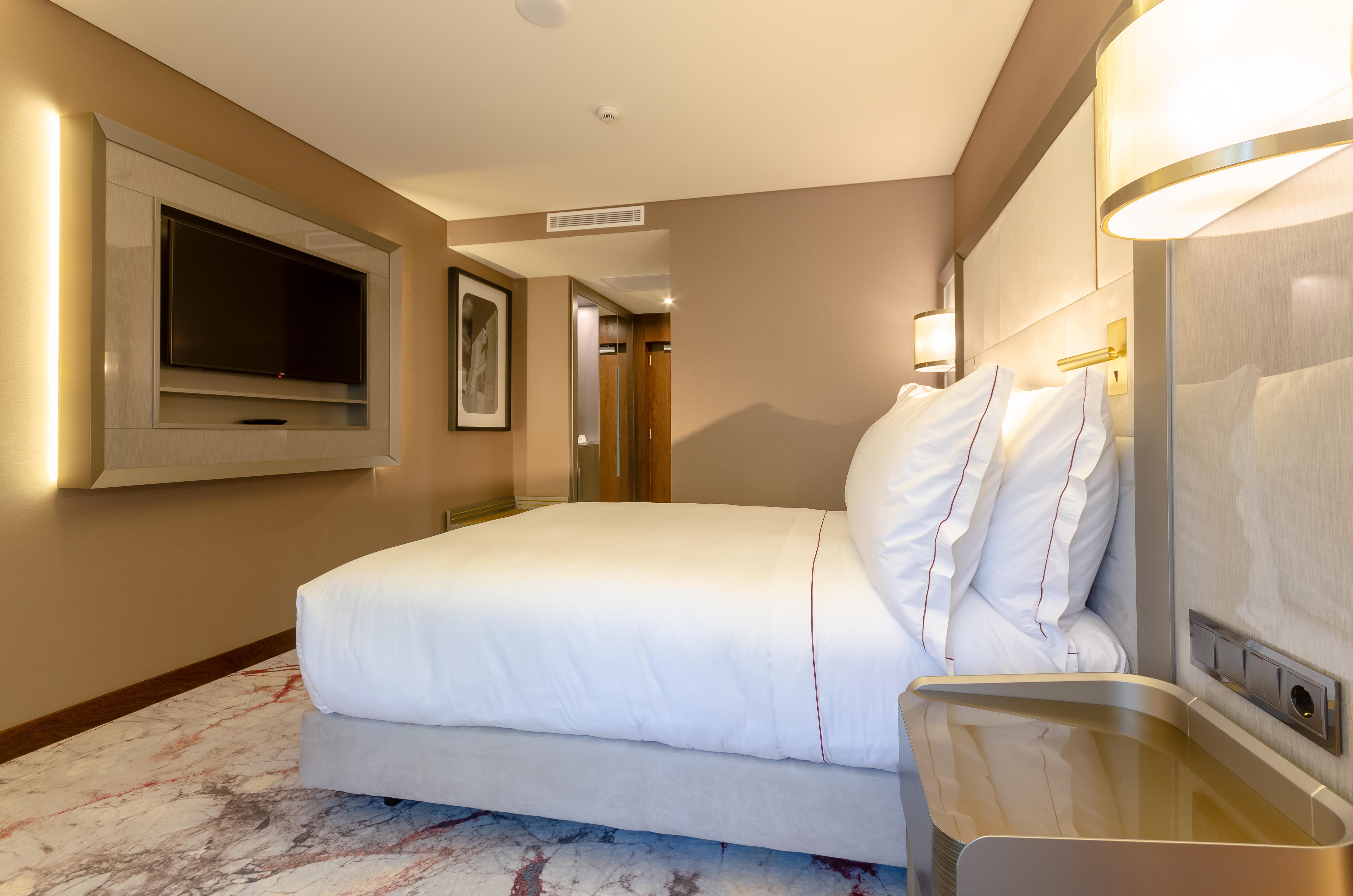 Turim Boulevard Hotel (Adults Only) Lisbona Esterno foto The photo shows a hotel room featuring a large, neatly made bed with white bedding and multiple pillows. The room has neutral-colored walls with modern decor, including a wall-mounted television opposite the bed. A bedside table is located next to th