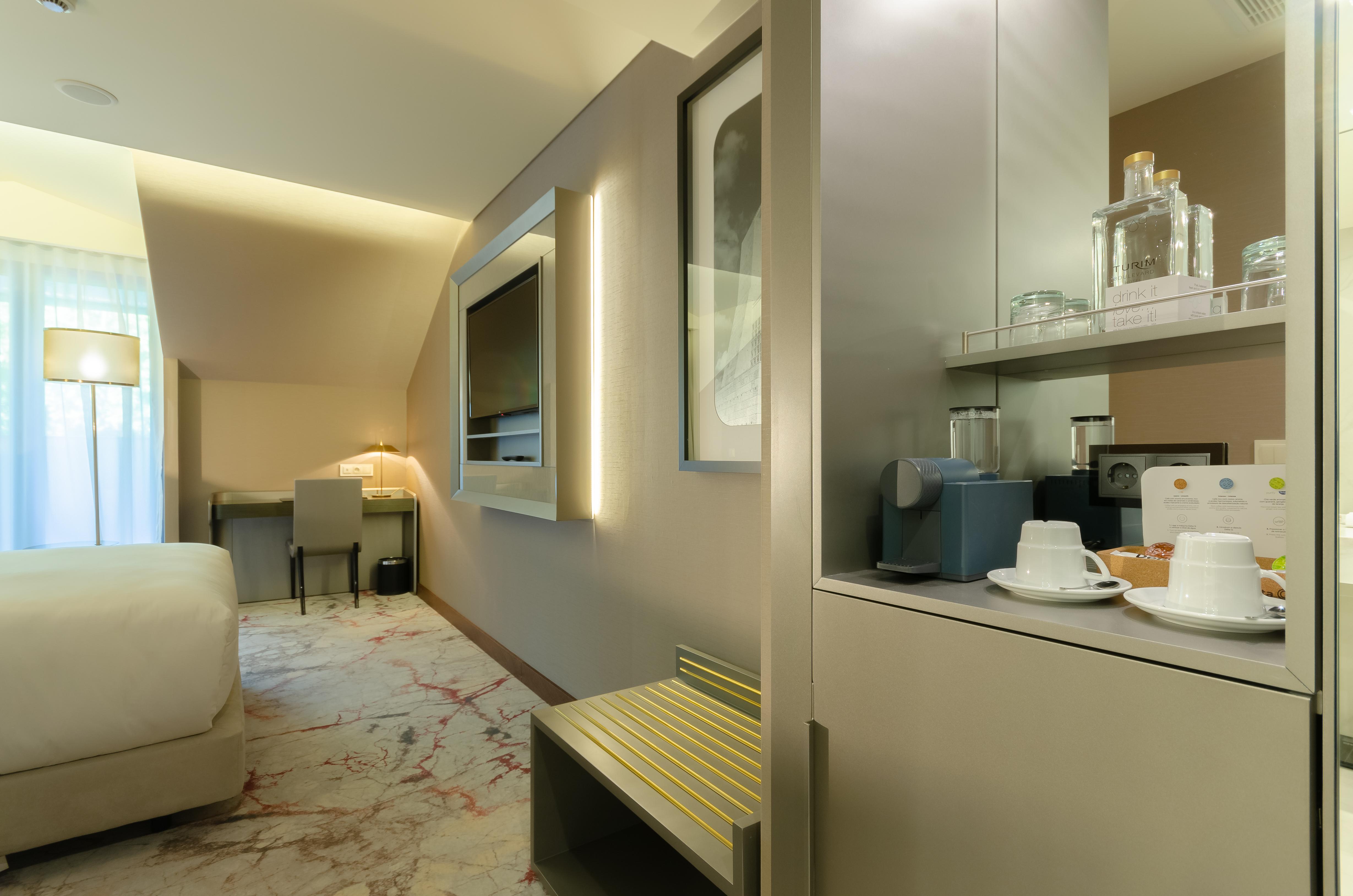 Turim Boulevard Hotel (Adults Only) Lisbona Esterno foto The photo shows a modern and stylish hotel room. There is a neatly made bed with white bedding in the foreground, and behind it, a screen or TV mounted on the wall. To the side, there is a small seating area with a desk and a lamp. On the right side 