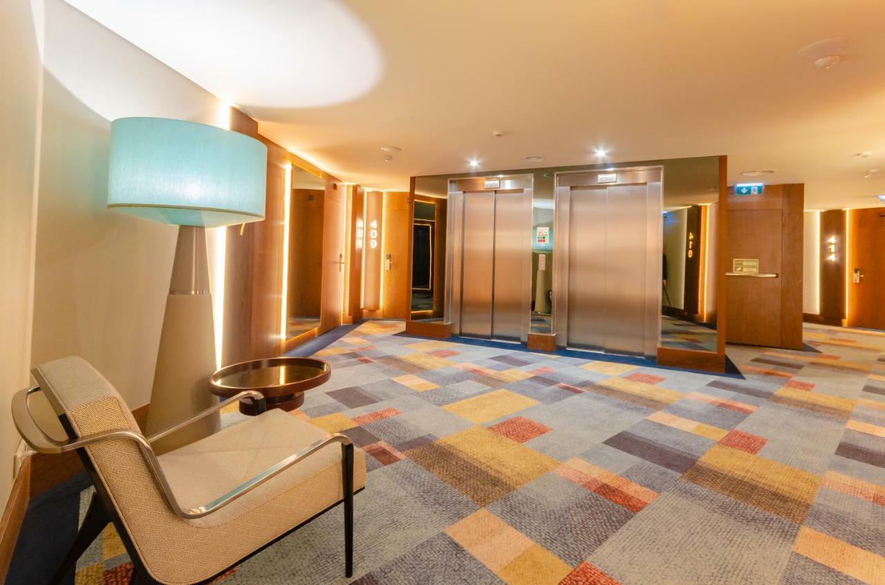 Turim Boulevard Hotel (Adults Only) Lisbona Esterno foto The photo shows a modern hallway presumably in a hotel or office building. On one side, there is a stylish light blue lamp next to a contemporary armchair. The floor is covered with a patterned carpet featuring squares of various colors, including bl