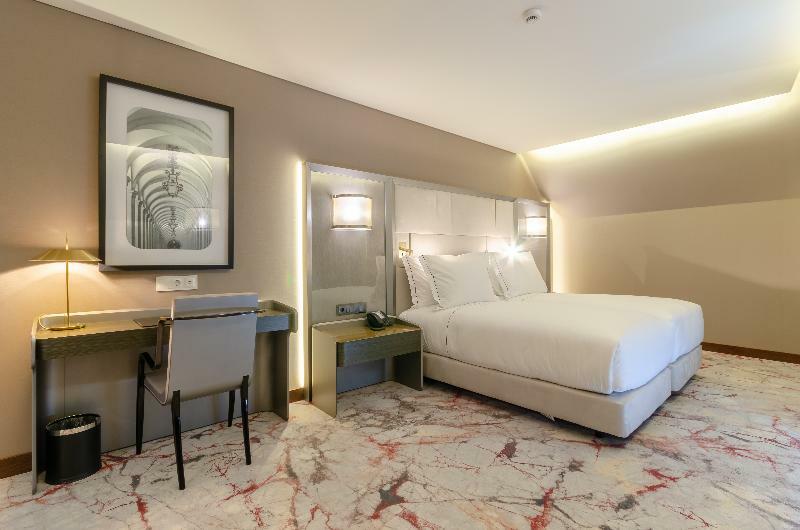 Turim Boulevard Hotel (Adults Only) Lisbona Esterno foto The photo shows a modern hotel room featuring a large bed with white linens against a neutral-colored wall. On one side of the bed, there are stylish wall-mounted lights. There is a small desk with a chair next to it, and a telephone is placed on the