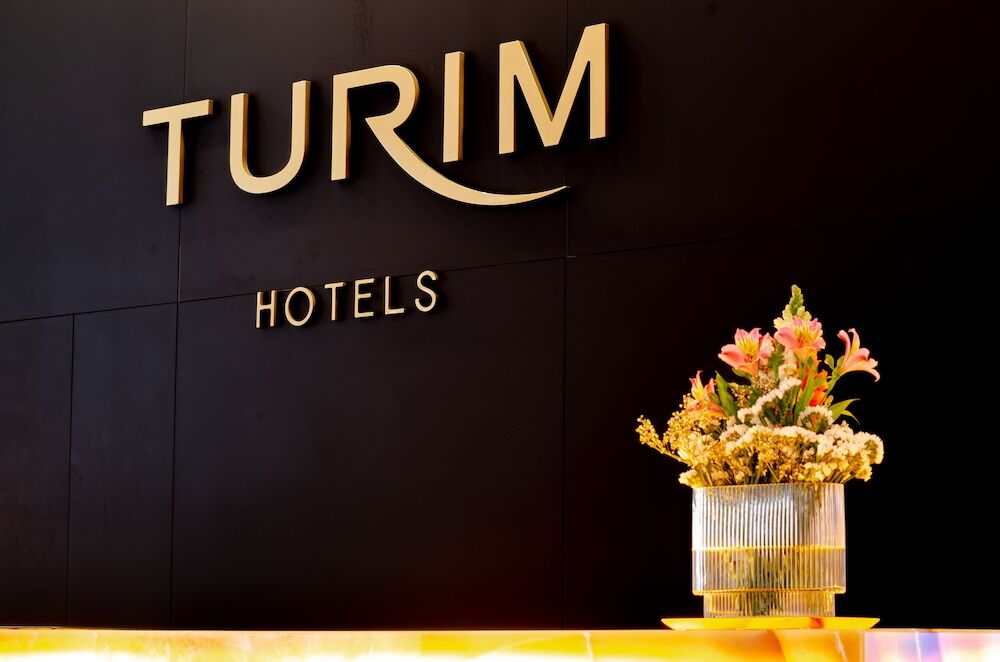 Turim Boulevard Hotel (Adults Only) Lisbona Esterno foto The photo shows a stylish reception area with the name "TURIM HOTELS" prominently displayed in gold lettering on a dark wall. Below the sign, there is a decorative arrangement of flowers in a sleek, modern vase. The overall atmosphere appears elegant