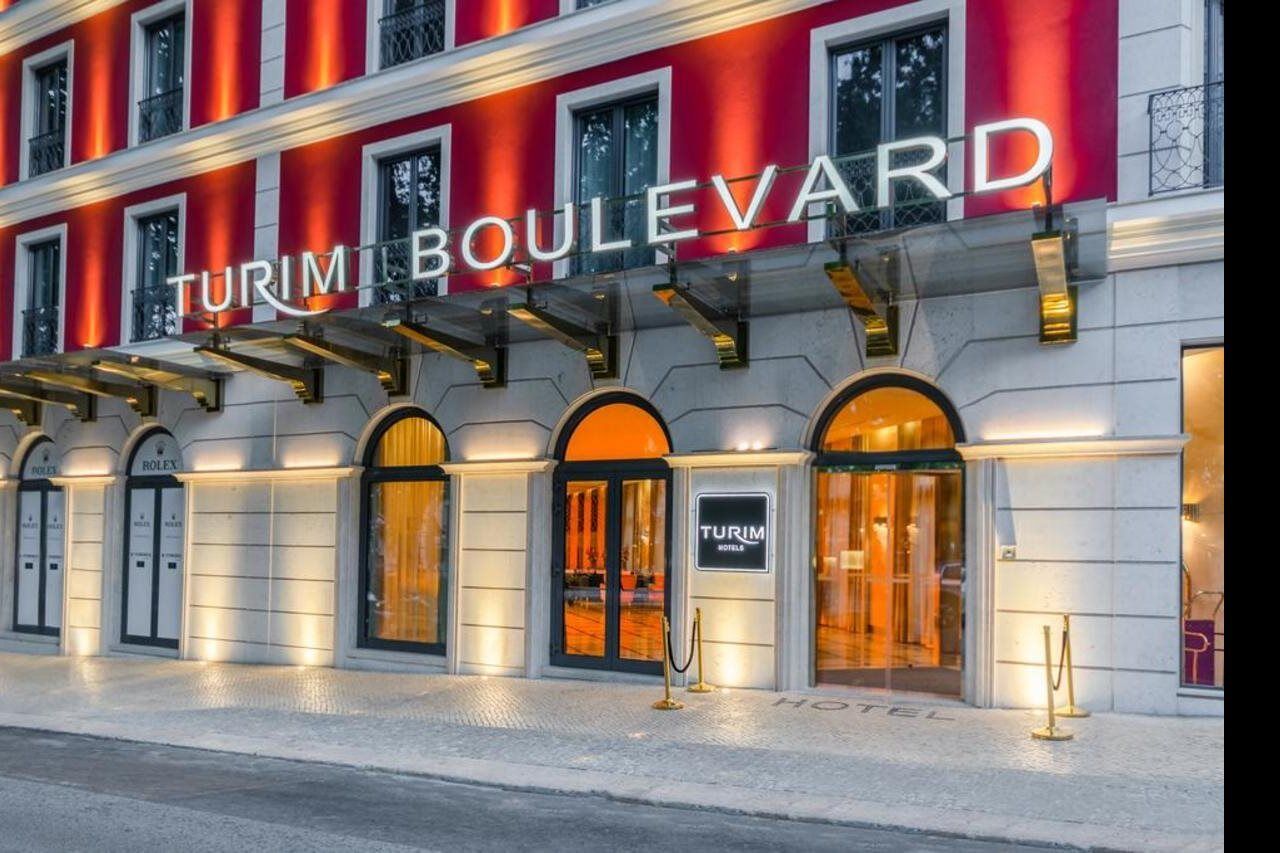 Turim Boulevard Hotel (Adults Only) Lisbona Esterno foto The photo shows the exterior of a hotel named "Turim Boulevard." The building has a modern design with a bright facade featuring red accents. Large windows are arched, enhancing the architectural style. There is illuminated signage with the hotel's n